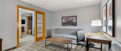 A hotel room with a sofa, a coffee table, a desk with a lamp, a painting on the wall, and a checkered carpet.