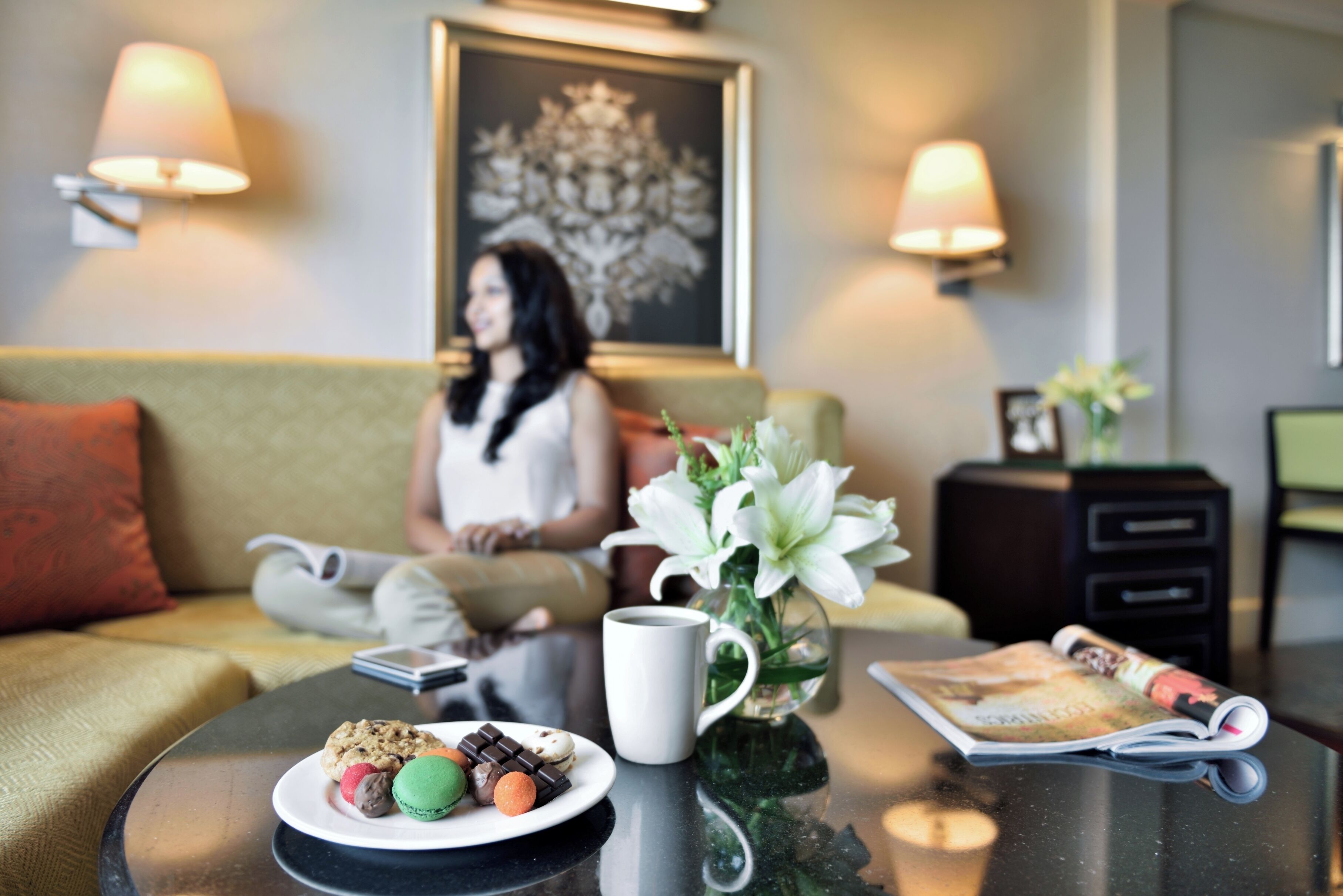 A modern hotel room with a sofa, a coffee table with pastries, a magazine, and a vase with flowers.