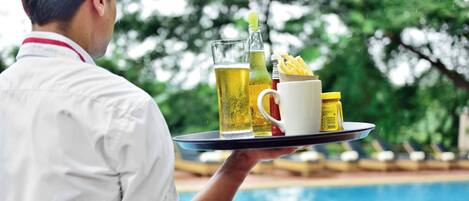 Une personne tenant un plateau sur lequel se trouvent un verre de bière, une bouteille, une tasse et des condiments, près d’une piscine.