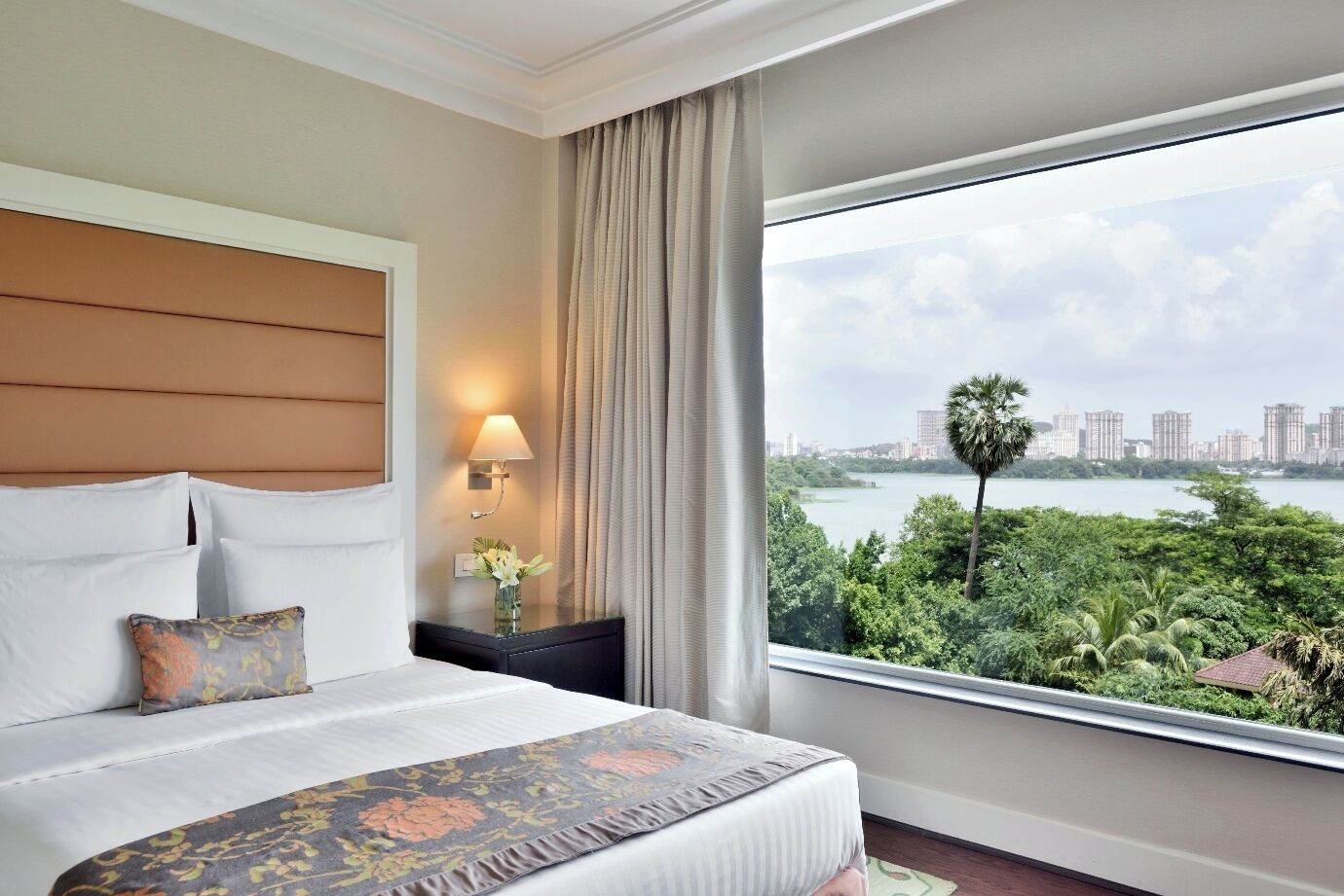 A hotel room with a bed, a nightstand with a lamp, a vase with flowers, and a large window offering a view of a city and greenery.
