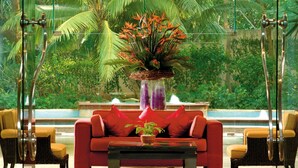 Um sofá vermelho com um vaso de planta em frente, rodeado por cadeiras de vime, com vista para uma fonte e vegetação exuberante.