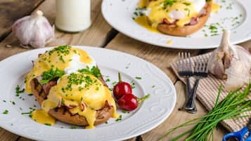 A breakfast setting with eggs Benedict on white plates, garnished with chives, and surrounded by garlic bulbs and chives.