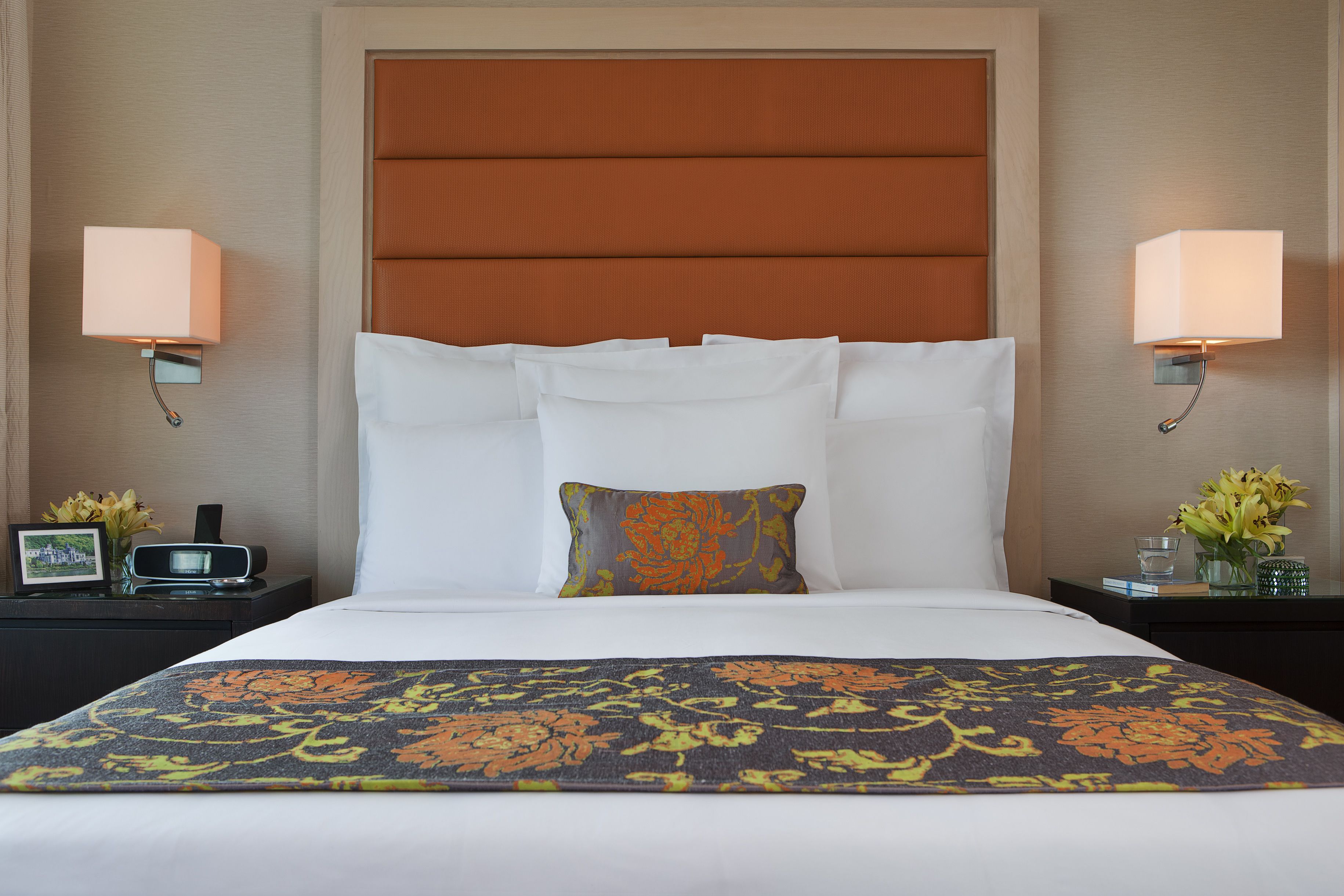 Habitación de hotel con una cama grande, dos mesitas de noche, una mesita auxiliar, un cabecero y dos lámparas fijadas a la pared.