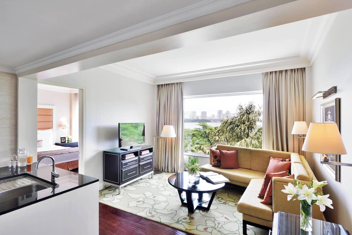 A modern hotel room with a kitchen area, a living room with a sofa and a coffee table, and a large window offering a city view.