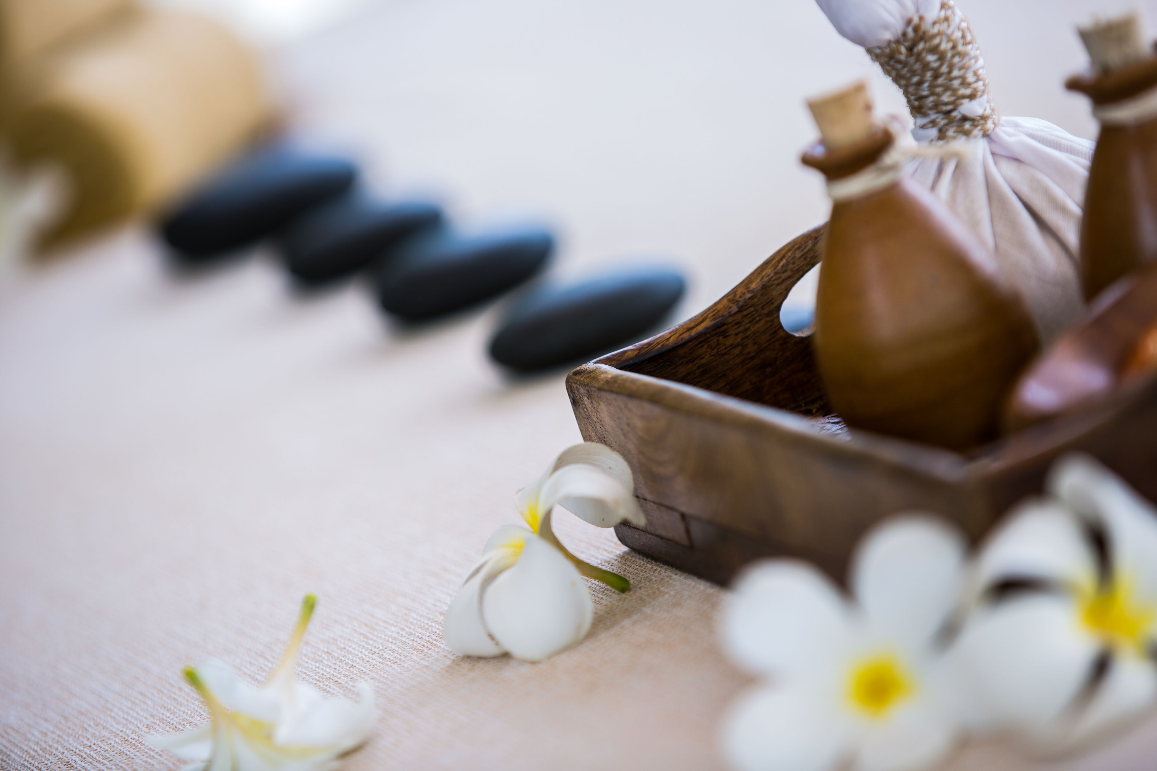 A wooden tray with a small bottle and a cloth, surrounded by white flowers and black stones on a textured surface.