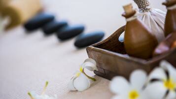 A wooden tray with a small bottle and a cloth, surrounded by white flowers and black stones on a textured surface.