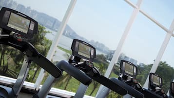 Drei Fitnessräder mit digitalen Anzeigen, platziert in einem Raum mit großen Fenstern und Blick auf Wasser und Grünflächen.