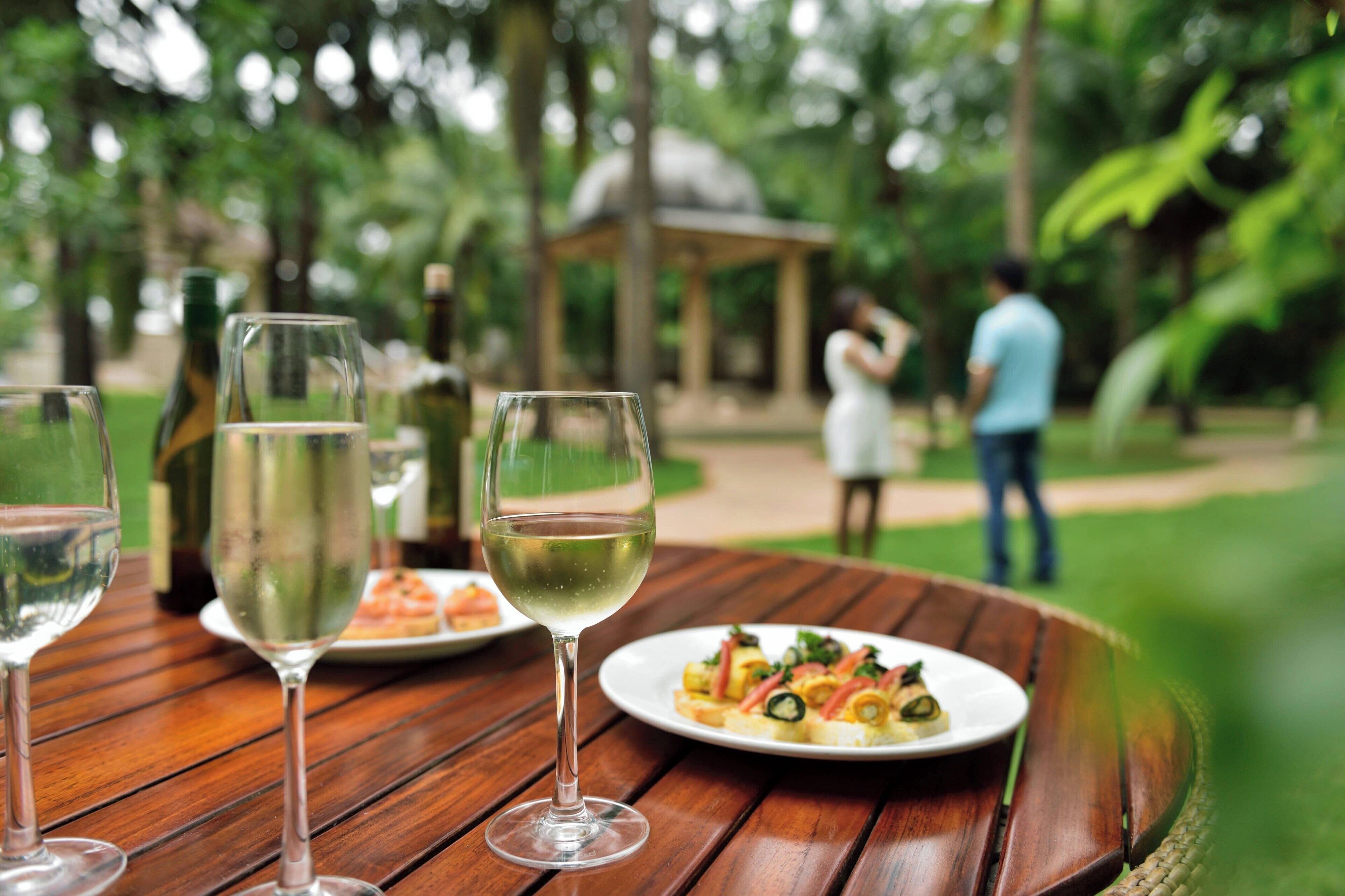 Une table en bois avec trois verres à vin et une assiette de nourriture, le tout installé à l’extérieur, avec de la verdure en arrière-plan.