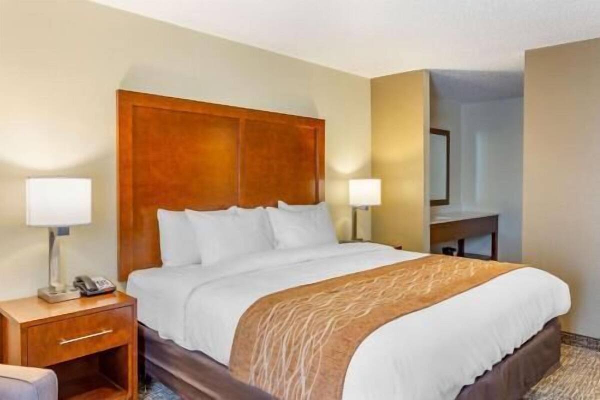 A hotel room with a wooden headboard, a bed with white bedding, two bedside lamps, a nightstand with a phone, and a mirror on the wall.