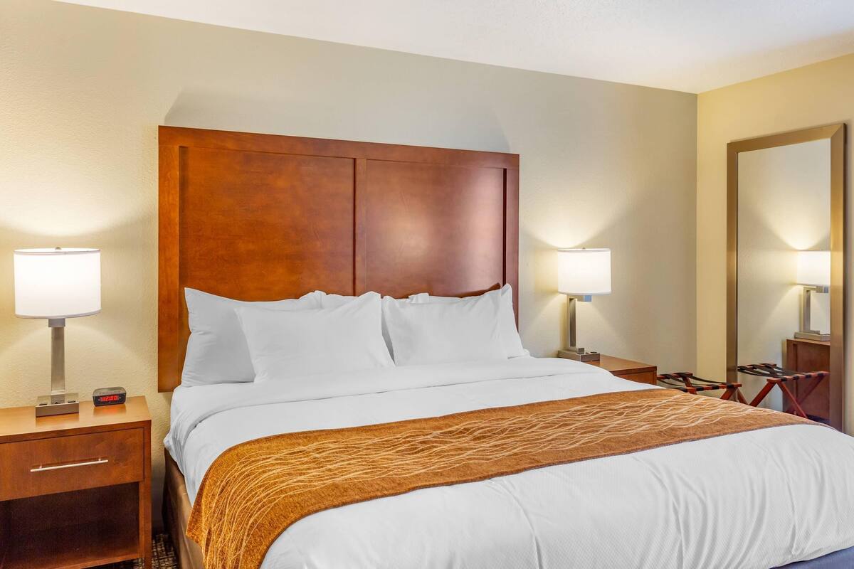 A hotel room with a wooden headboard, white bedding, two bedside lamps, a clock, and a large mirror.