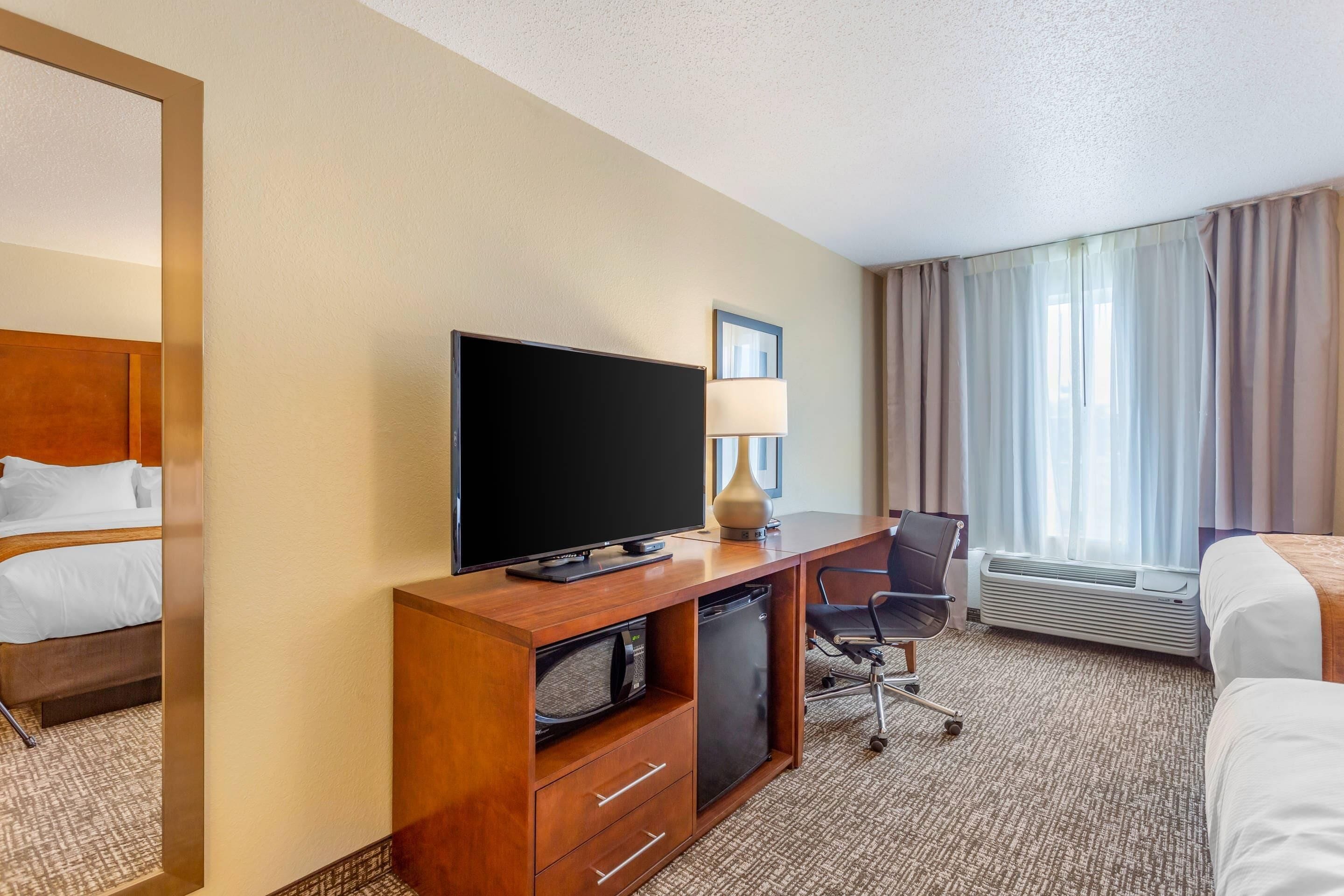 Habitación de hotel con cama, escritorio con televisor, silla, microondas, refrigerador y una ventana grande con cortinas.