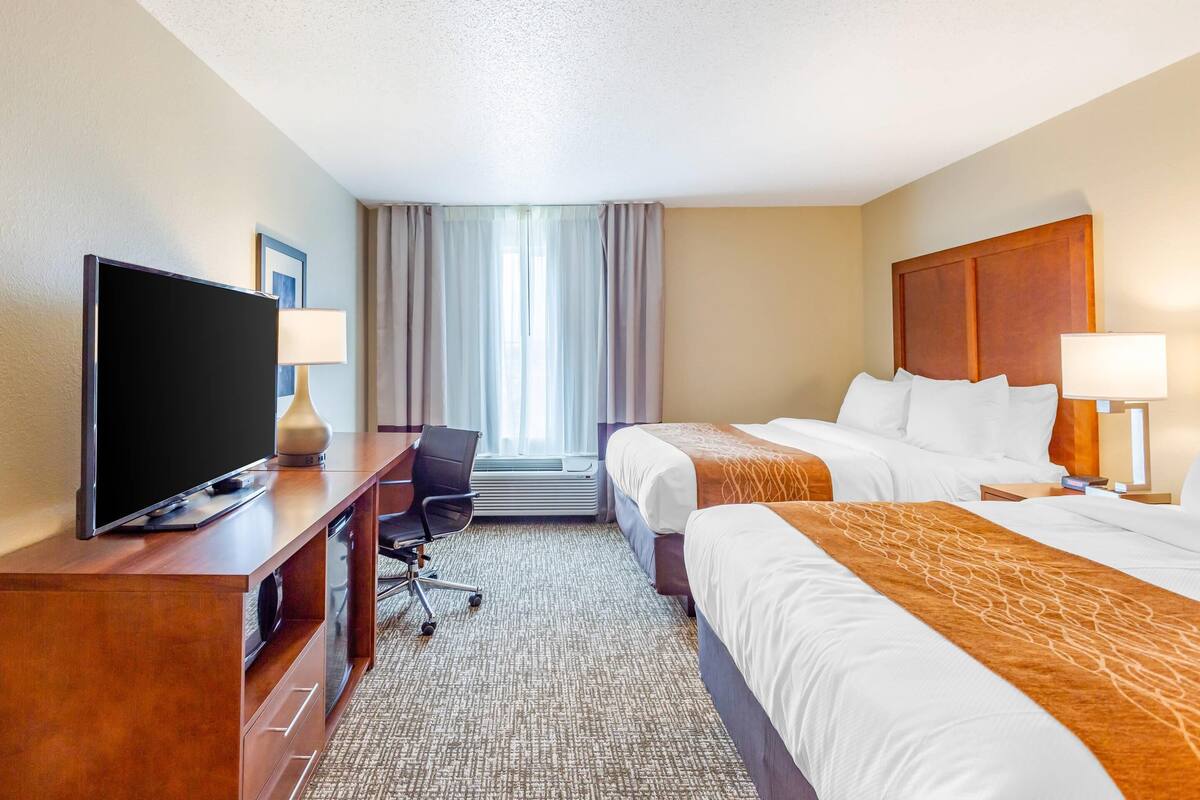 A hotel room with a TV, desk, two beds, and a window with curtains.