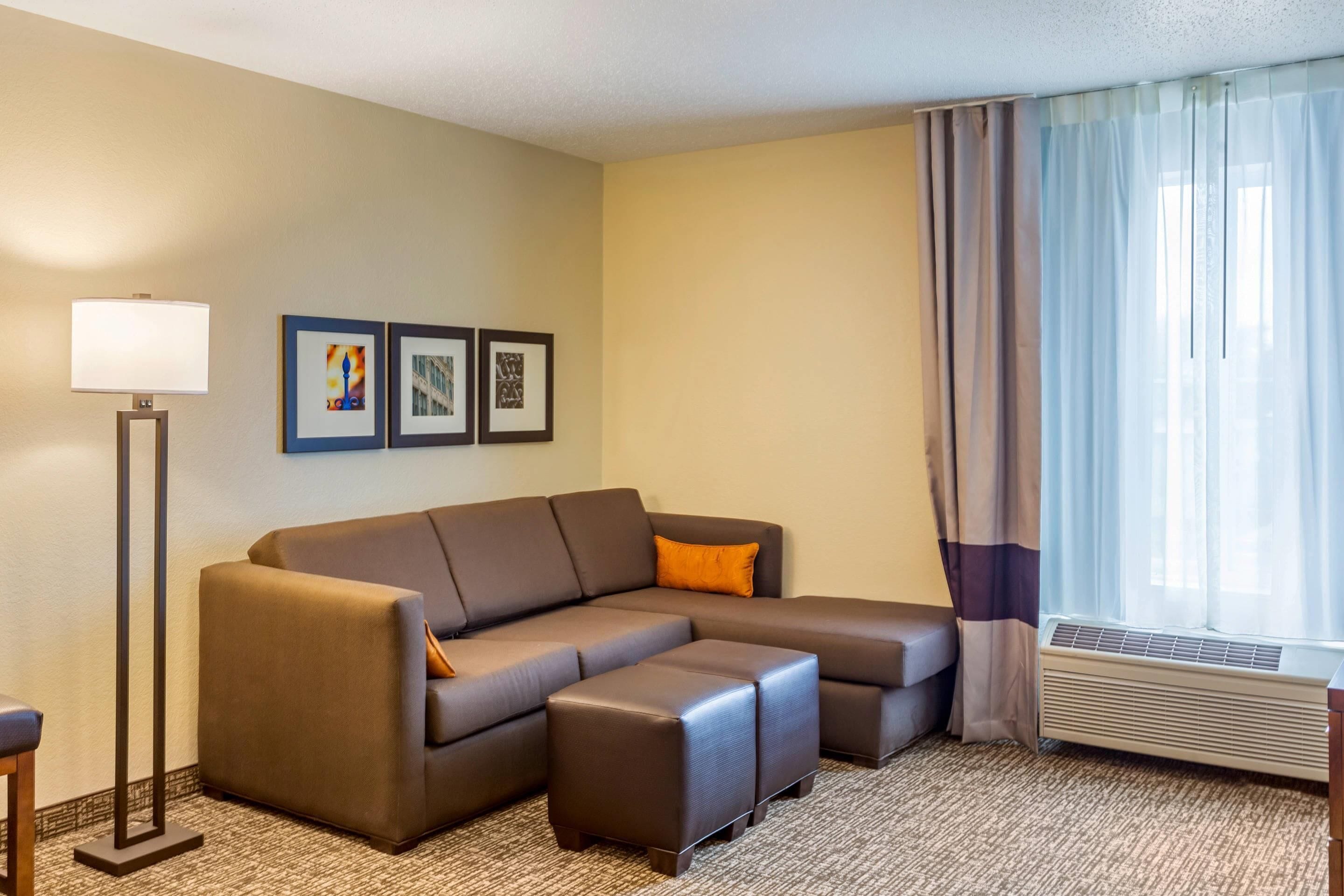 A modern hotel room with a sectional sofa, a standing floor lamp, framed artwork on the wall, and a window with curtains.