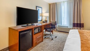 Una camera d'albergo con televisione a schermo piatto, una scrivania con lampada, una sedia, un letto e una finestra con tende.