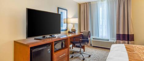 Habitación de hotel con televisión de pantalla plana, escritorio con lámpara, una silla, cama y ventana con cortinas.
