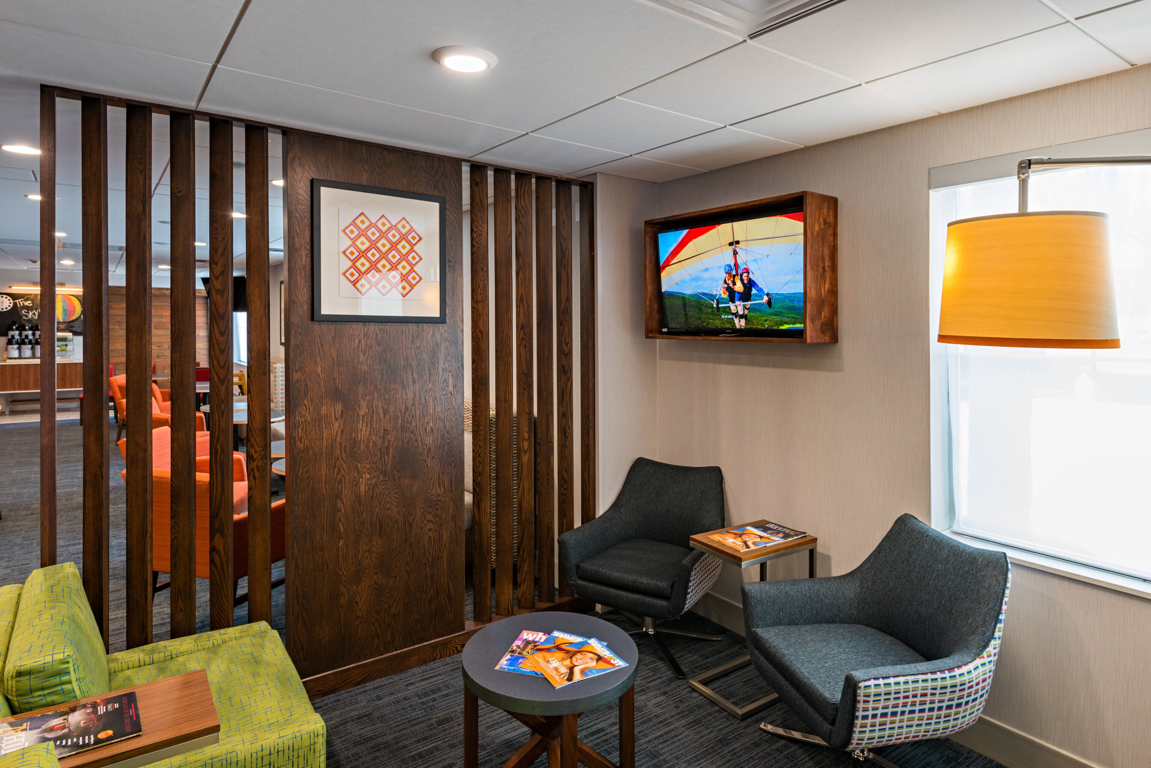 A hotel room with a wooden partition, a television mounted on the wall, a window with a lamp, and two armchairs.