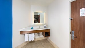A modern bathroom with a white sink, mirror, and wooden vanity. There are towels hanging on the wall and a door with a wooden panel. - Holiday Inn Express Hotel & Suites PEORIA NORTH - GLENDALE by IHG (Peoria)
