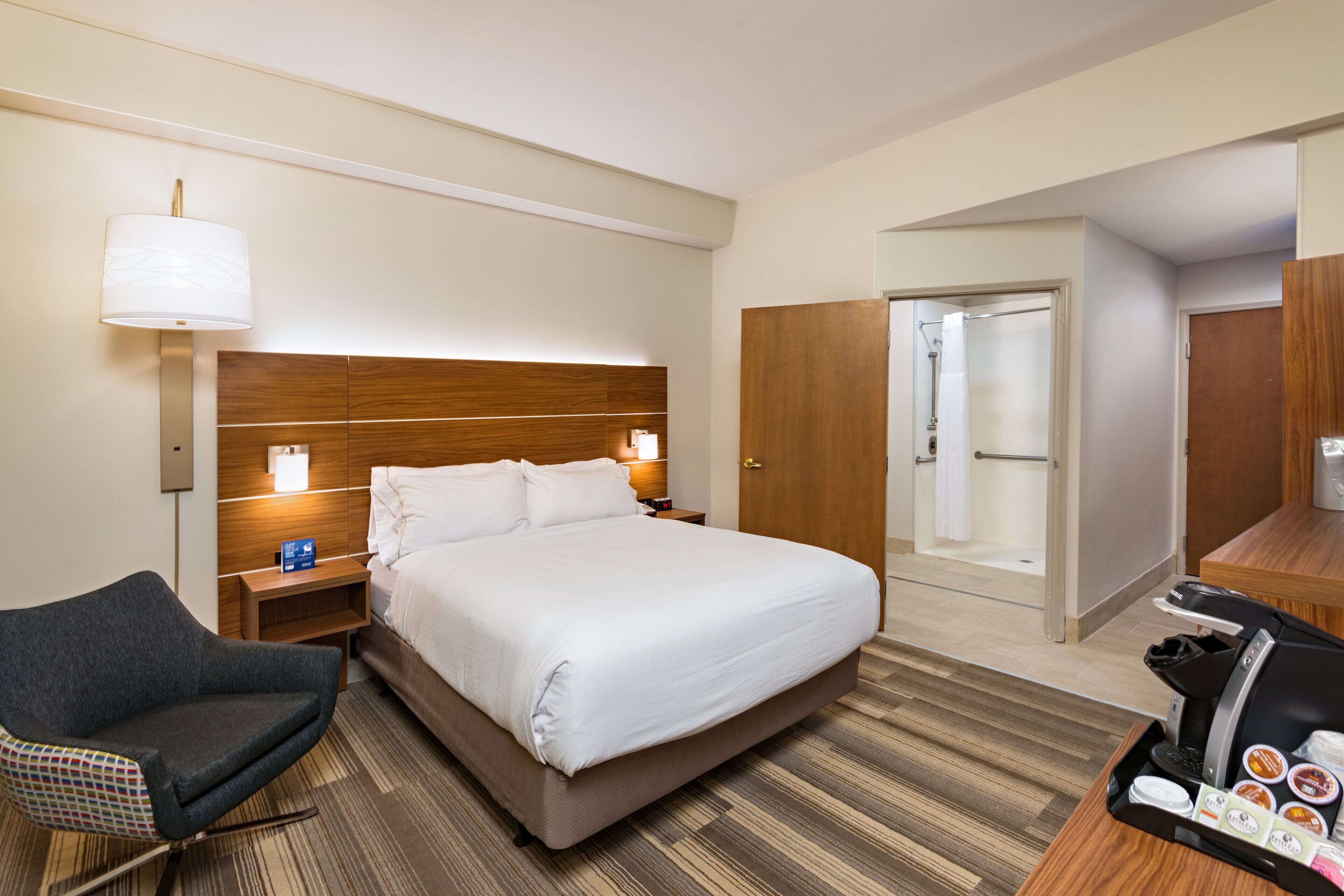 A hotel room with a bed, bedside tables, a chair, a desk with a coffee machine, and a bathroom with a shower and toilet.