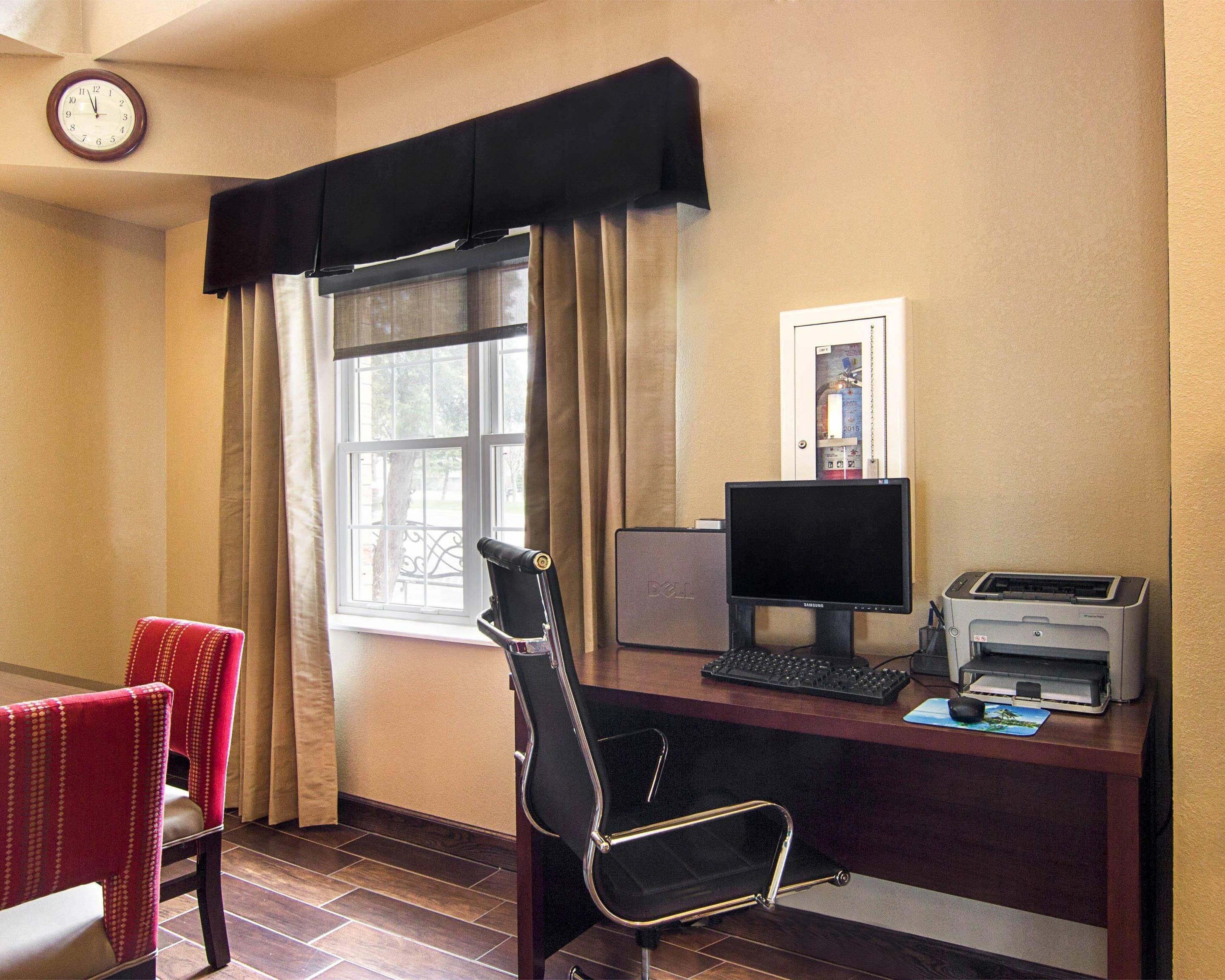 A hotel room with a desk, computer, printer, and a clock on the wall.