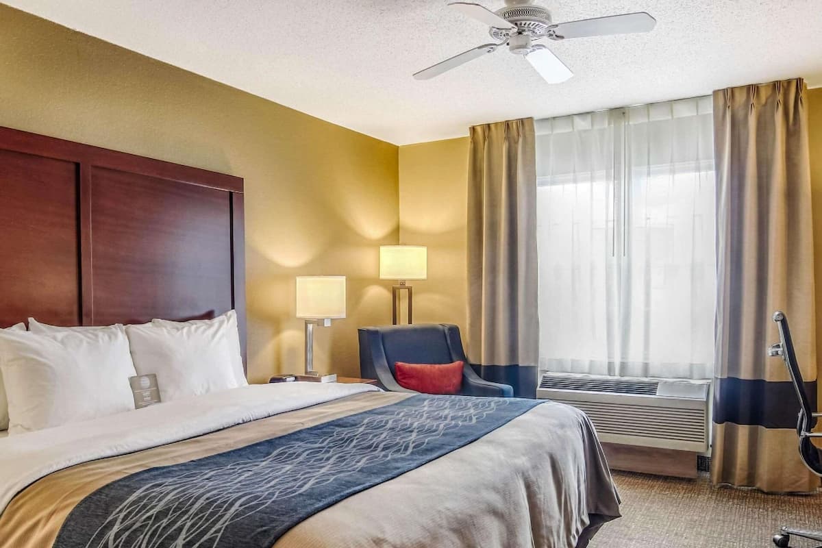 Une chambre d’hôtel comprenant un lit, un bureau, une chaise, une lampe de bureau, un ventilateur de plafond et une fenêtre avec des rideaux.