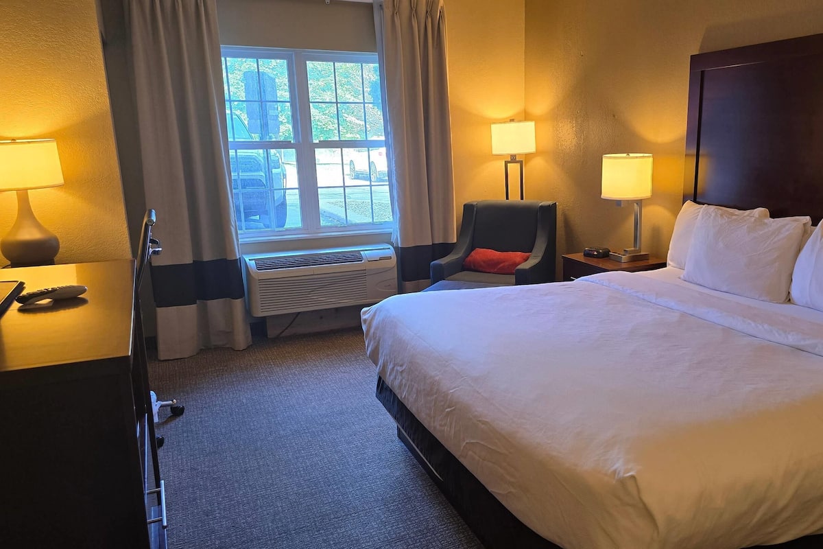 Une chambre d’hôtel avec un grand lit, une chaise, un bureau avec une lampe et un appareil de climatisation.
