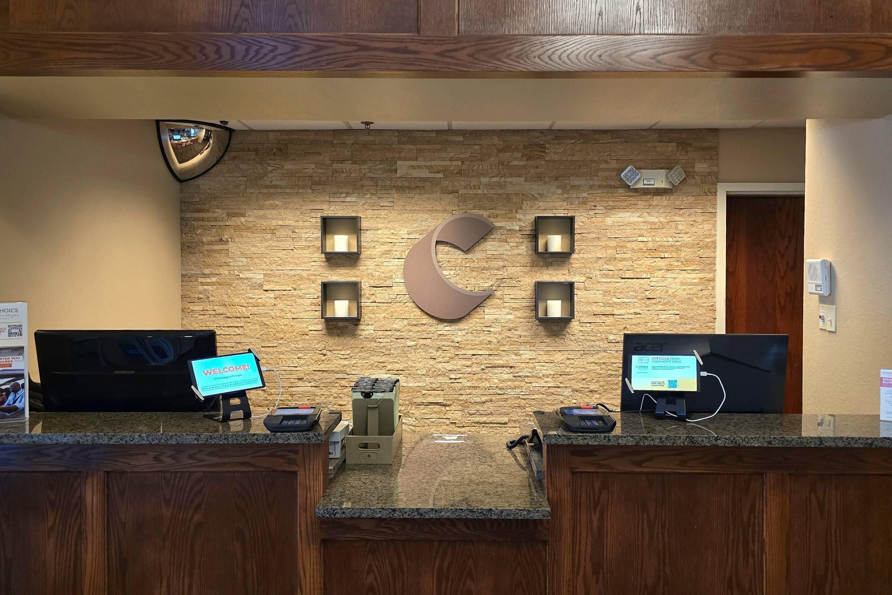 Un mostrador de recepción con un fondo de pared de piedra, un gran logotipo C, dos monitores, una máquina de café y un cartel de bienvenida.