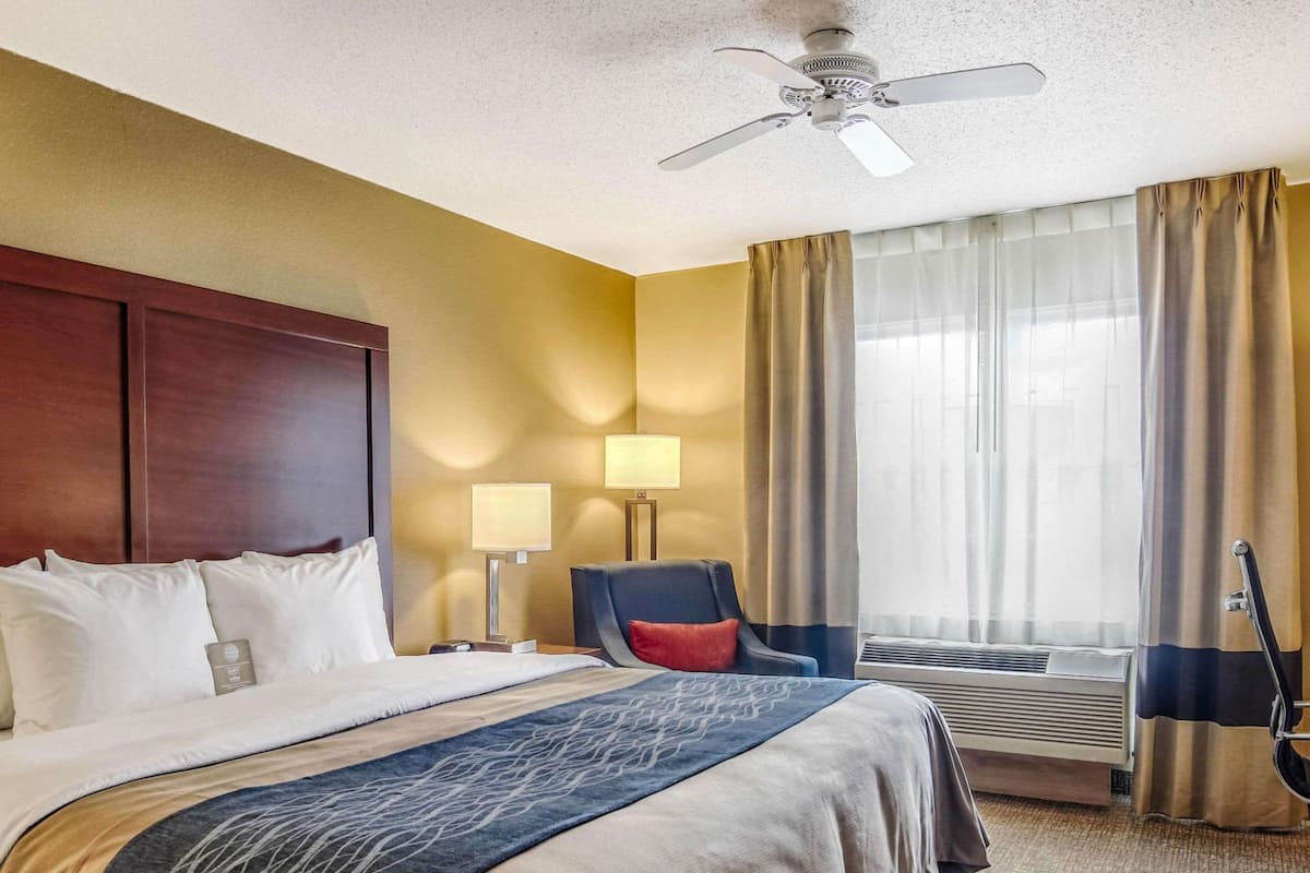 Une chambre d’hôtel avec un grand lit, un bureau, une chaise, une lampe de bureau, un ventilateur de plafond et une fenêtre avec des rideaux.