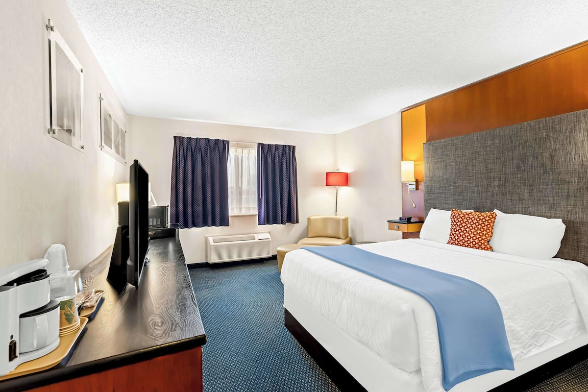 A hotel room with a bed, a desk with a coffee maker, a chair, a TV, and a window with curtains.