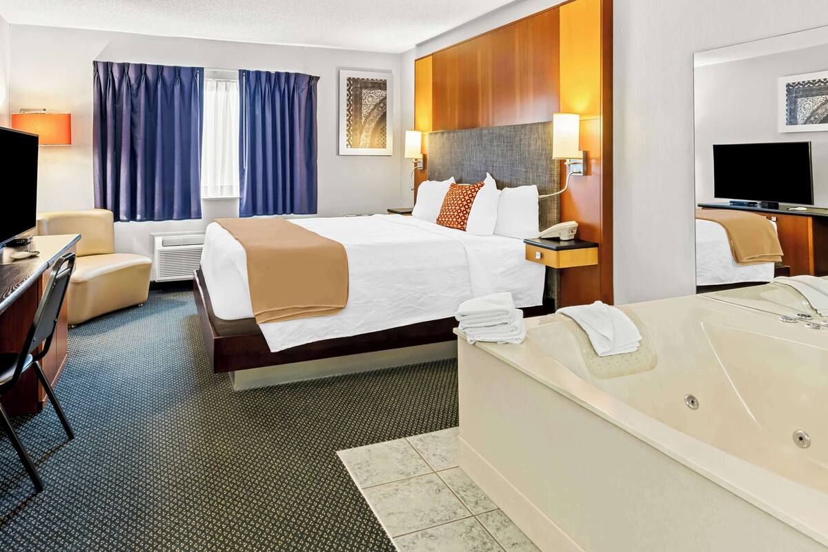 A hotel room with a large bed, a desk with a chair, a desk lamp, a television, a bathroom with a bathtub, and a window with curtains.