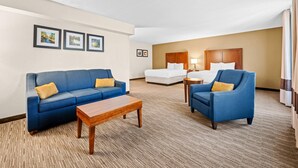 Camera d'albergo con un divano blu, un tavolino di legno, un letto bianco e una poltrona blu. Si notano quadri appesi al muro e una lampada sul comodino.
