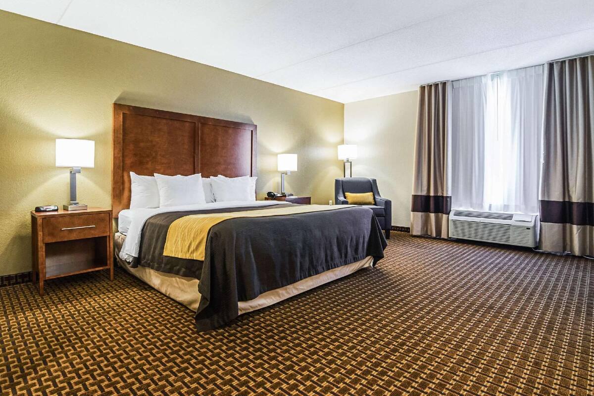 A hotel room with a bed, two bedside tables, a chair, a window with curtains, and an air conditioning unit.