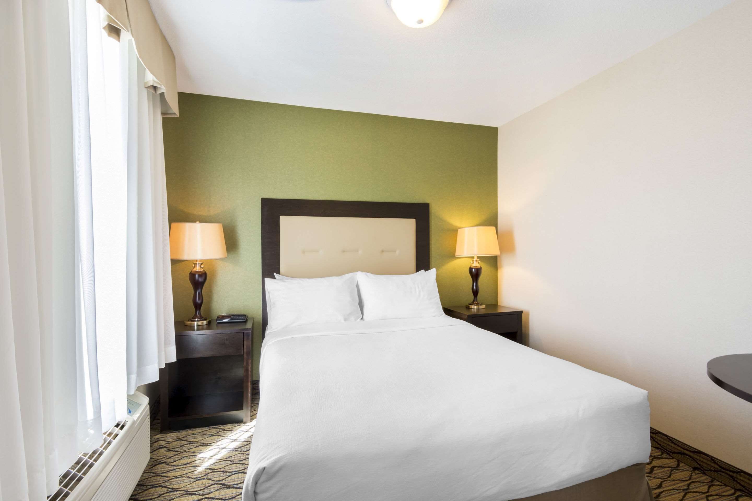 A hotel room with a bed, two bedside tables with lamps, a nightstand, a window with curtains, and a wall with a green accent.