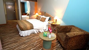 A hotel room with a bed, a sitting area with a chair, a small table with a vase of flowers, and a nightstand with a lamp. - La Vinci (Dhaka)