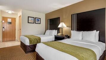 A hotel room with two beds, a wooden door, a nightstand with a lamp, and two framed pictures on the wall.