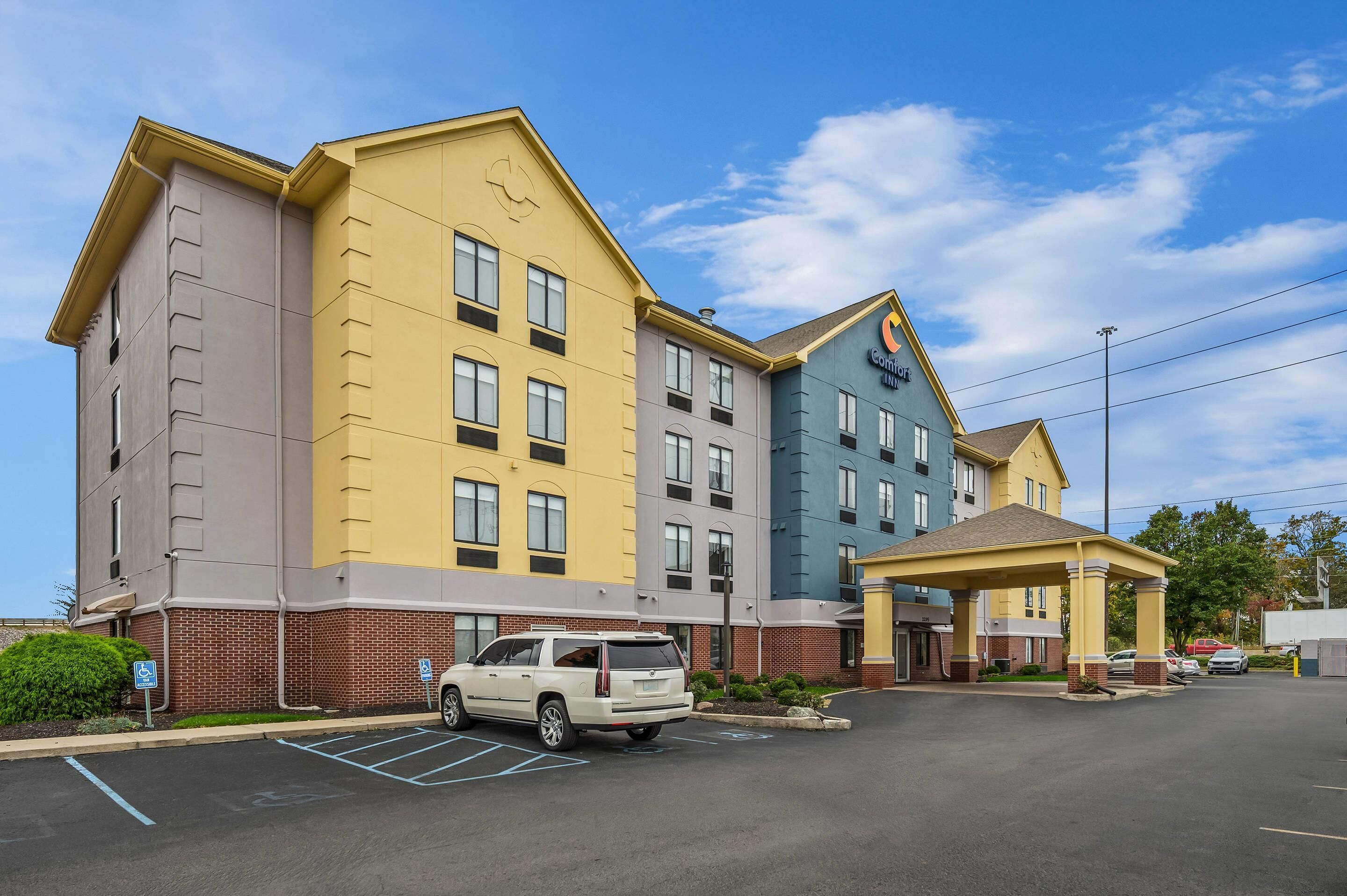 A multi-story hotel with a yellow and blue facade, a covered parking area, and a white SUV parked in the lot.