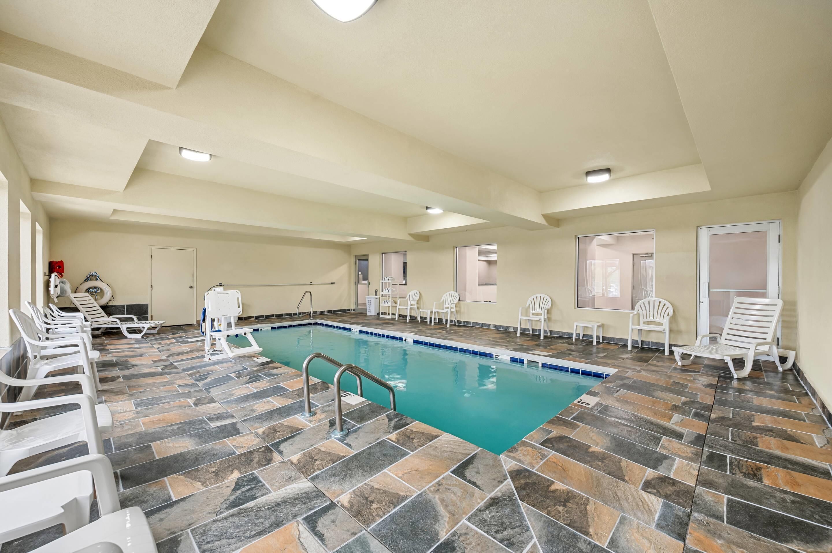 An indoor swimming pool with a tiled floor, white lounge chairs, and a ceiling with recessed lighting.