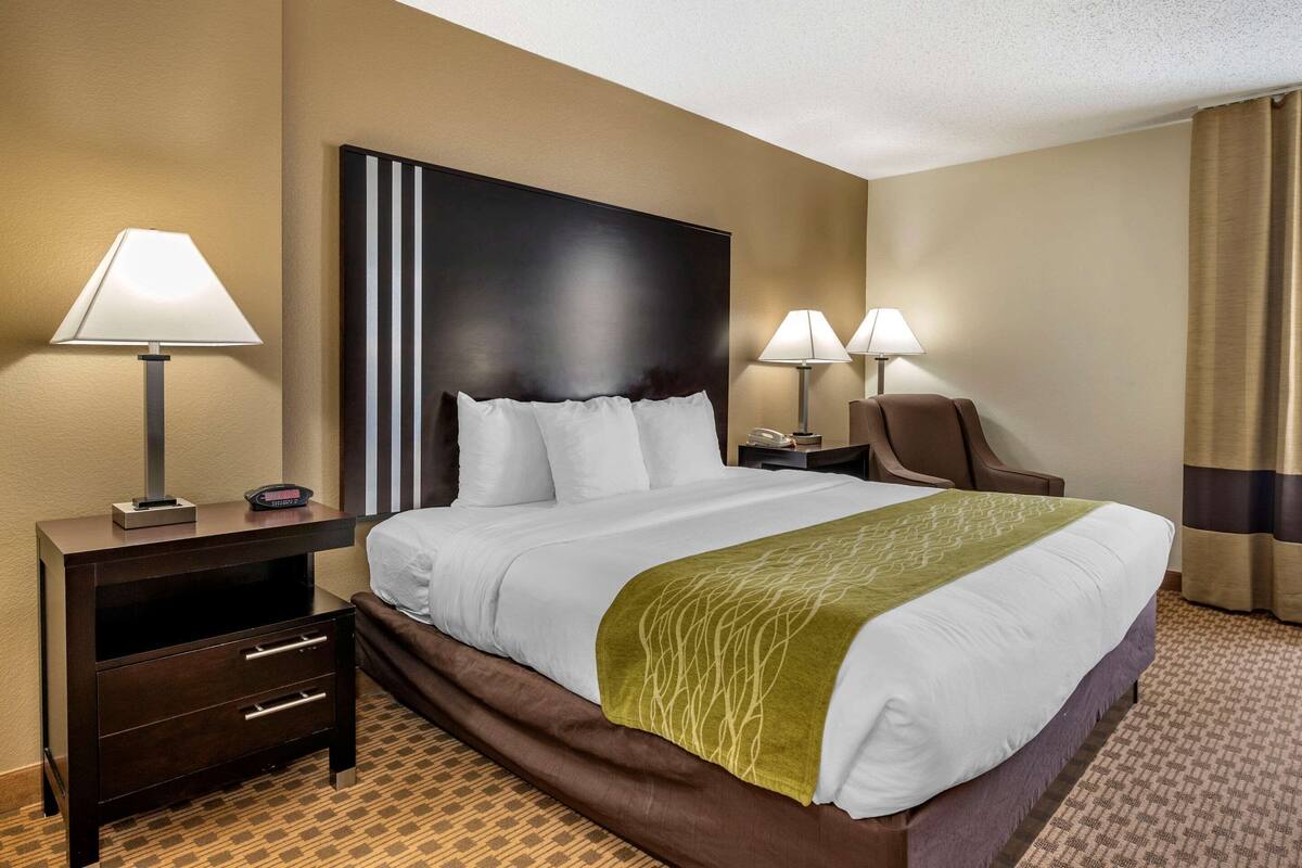 A hotel room with a large bed, two bedside tables with lamps, a chair, and a dark headboard.