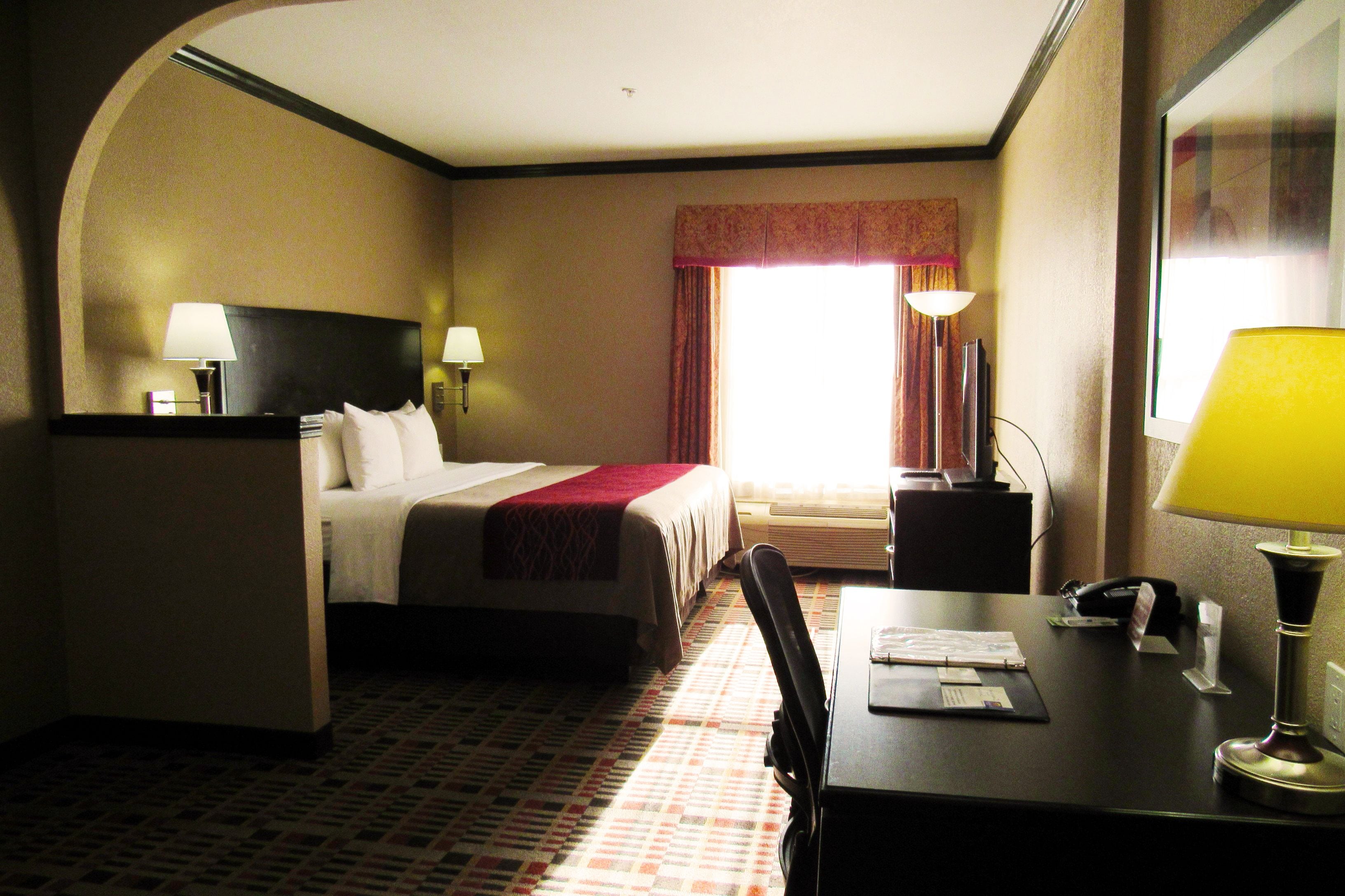 A hotel room with a bed, desk, chair, lamps, and a window with curtains.