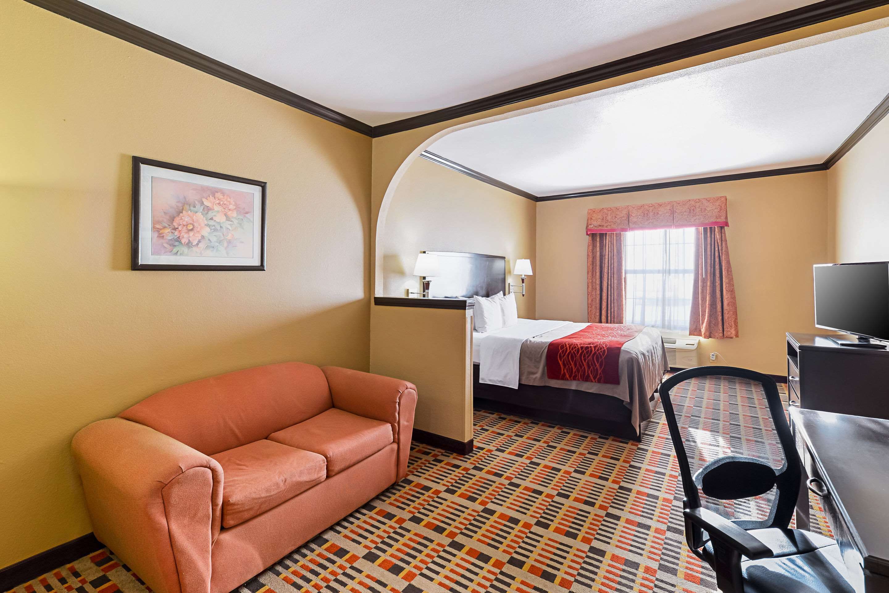 Une chambre d’hôtel avec un canapé, un lit recouvert d’une couverture rouge, un bureau avec une chaise et un téléviseur.