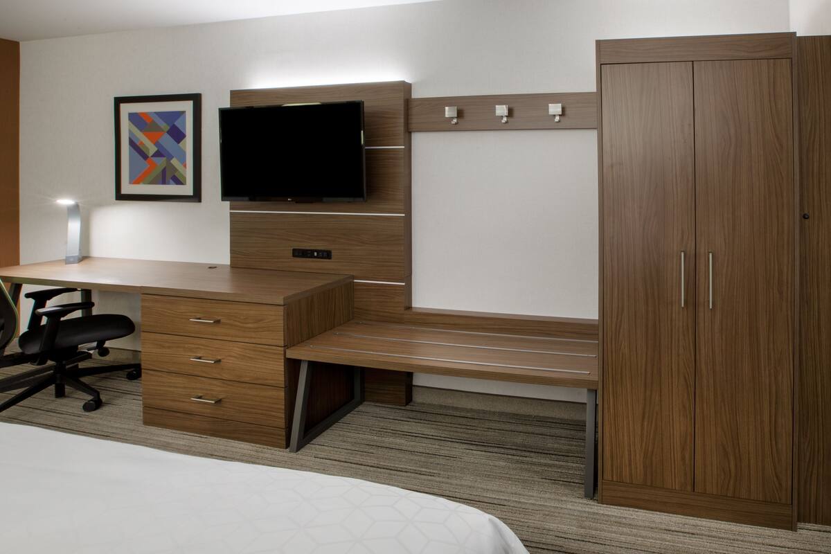 A hotel room with a wooden desk, a flat-screen TV, a bench, a wardrobe, and a bed.