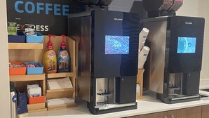 A coffee station with two espresso machines, labeled 'Costa Coffee', and a variety of coffee products and containers on shelves.