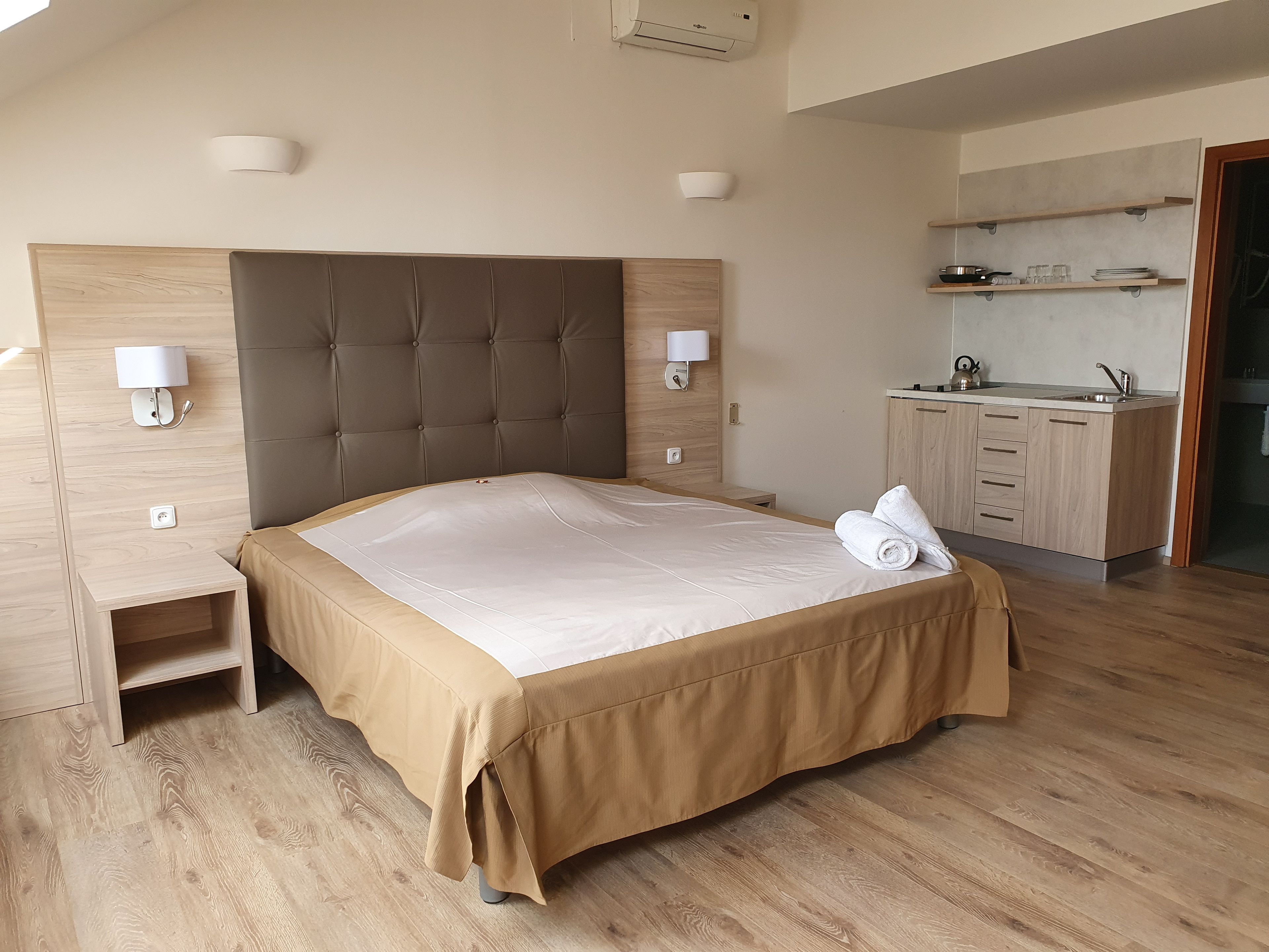 A modern hotel room with a large bed, a wooden headboard, a bedside table, a built-in kitchen area, and a view of the sky.