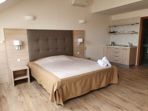 Ett modernt hotellrum med en stor säng, ett träbeklätt sänggavel, ett nattduksbord, ett inbyggt köksutrymme och utsikt över himlen.