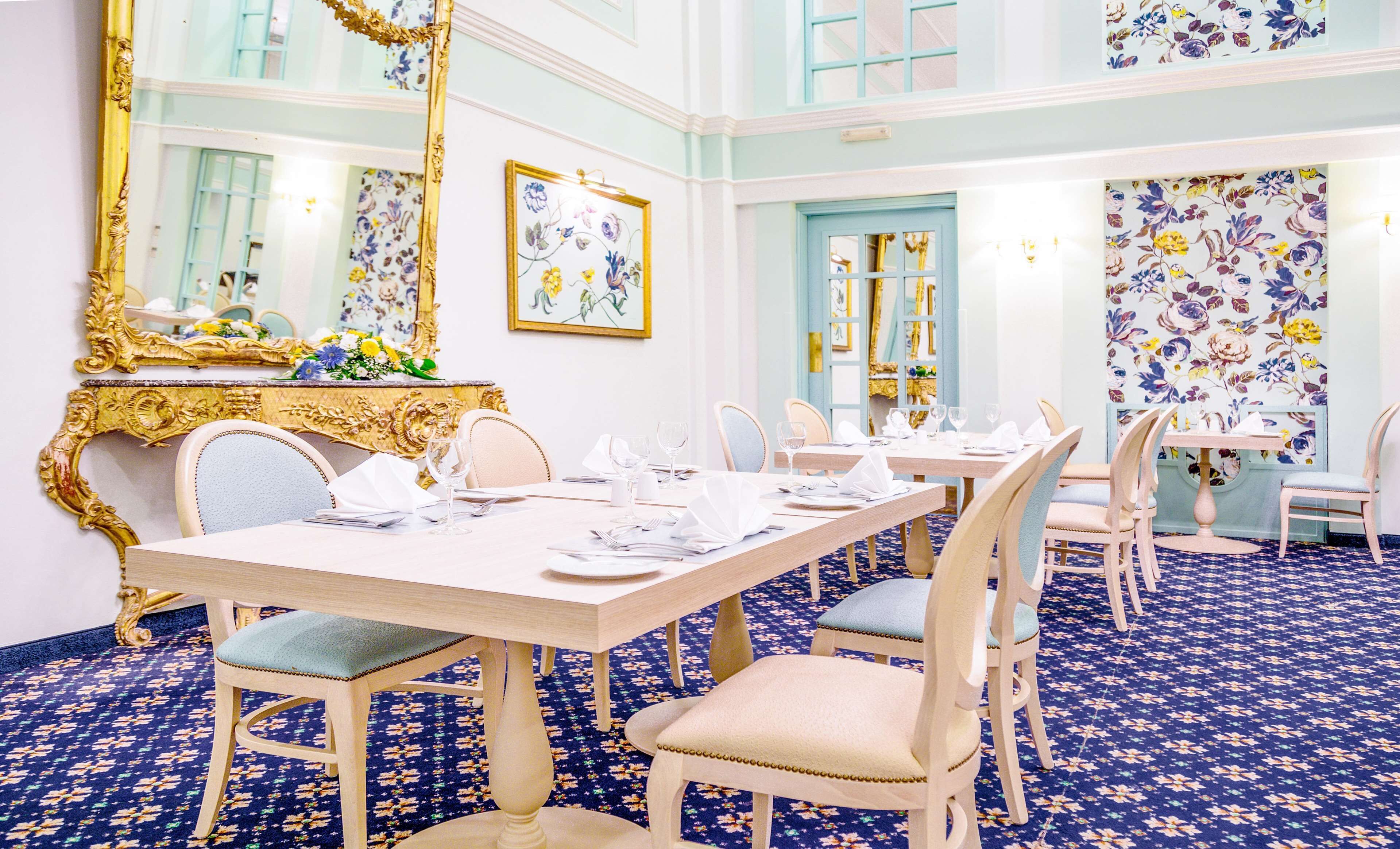 A dining area with a long table set for multiple guests, ornate gold-framed mirrors, floral wallpaper, and a patterned carpet.
