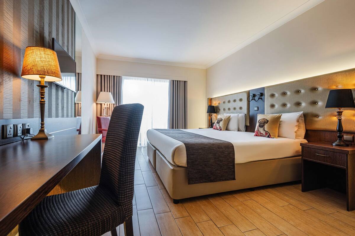 Une chambre d’hôtel avec un grand lit, un bureau avec une lampe, une chaise et une table de chevet avec une lampe.