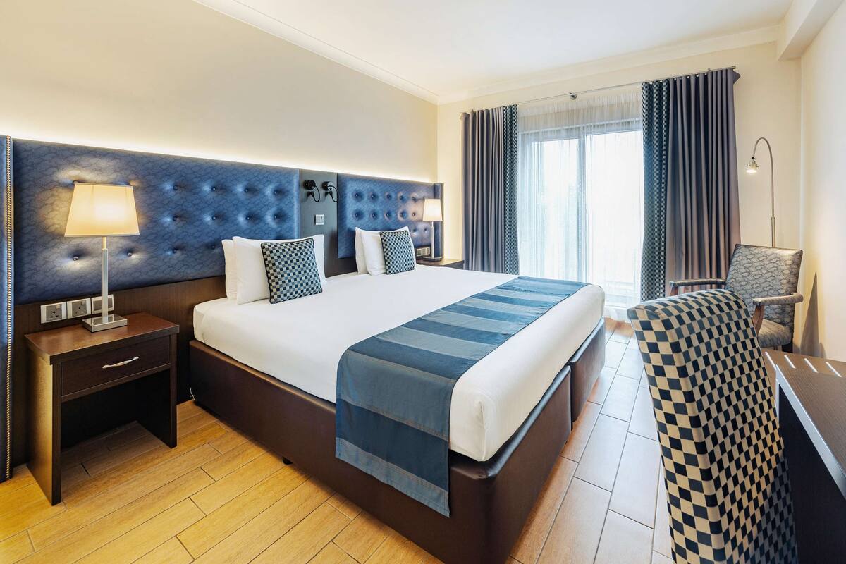 Une chambre d’hôtel avec un grand lit, une table de chevet avec une lampe, une chaise et une vue sur l’extérieur par la fenêtre.