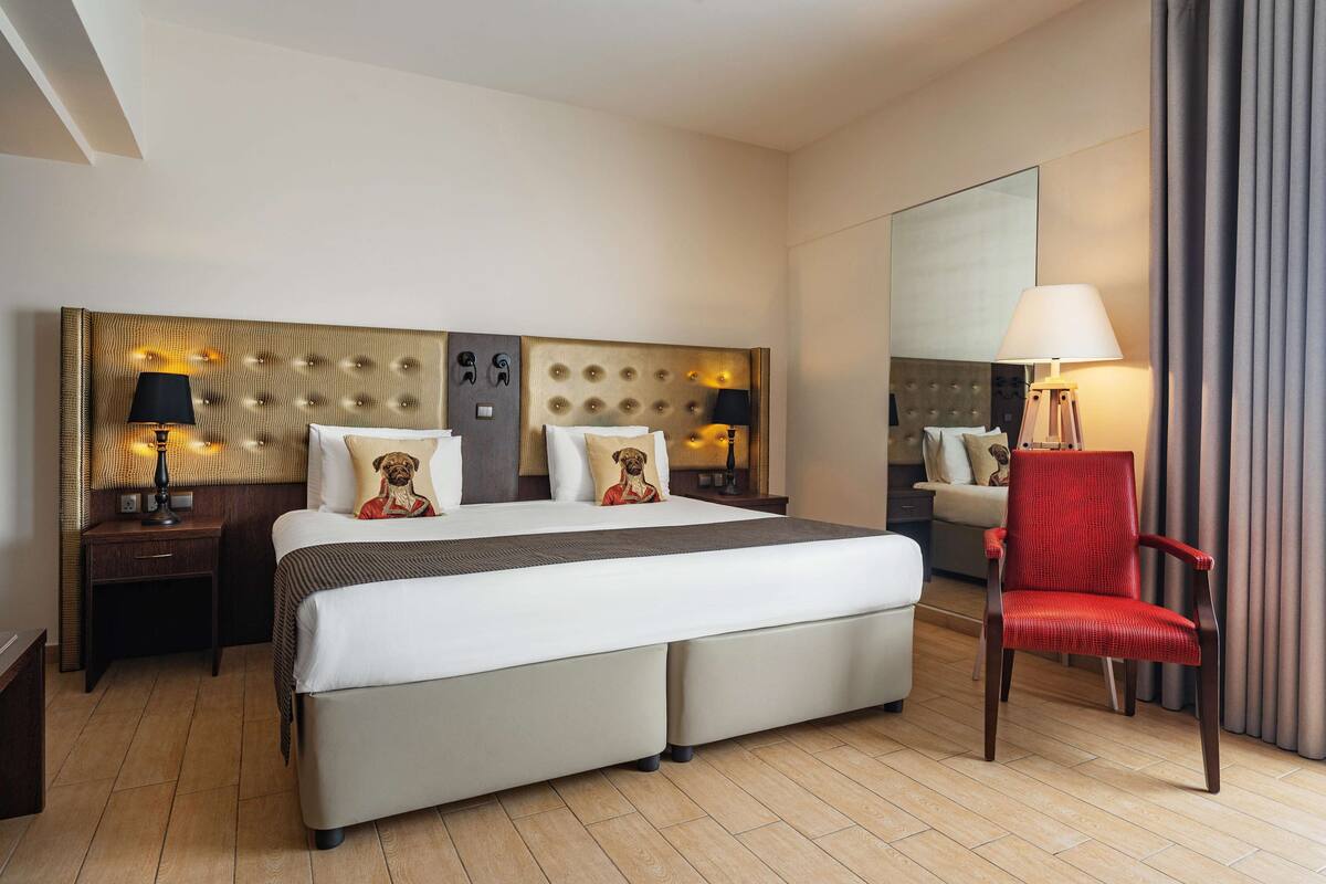 Une chambre d’hôtel avec un grand lit, deux tables de chevet avec des lampes, un fauteuil rouge et un grand miroir.