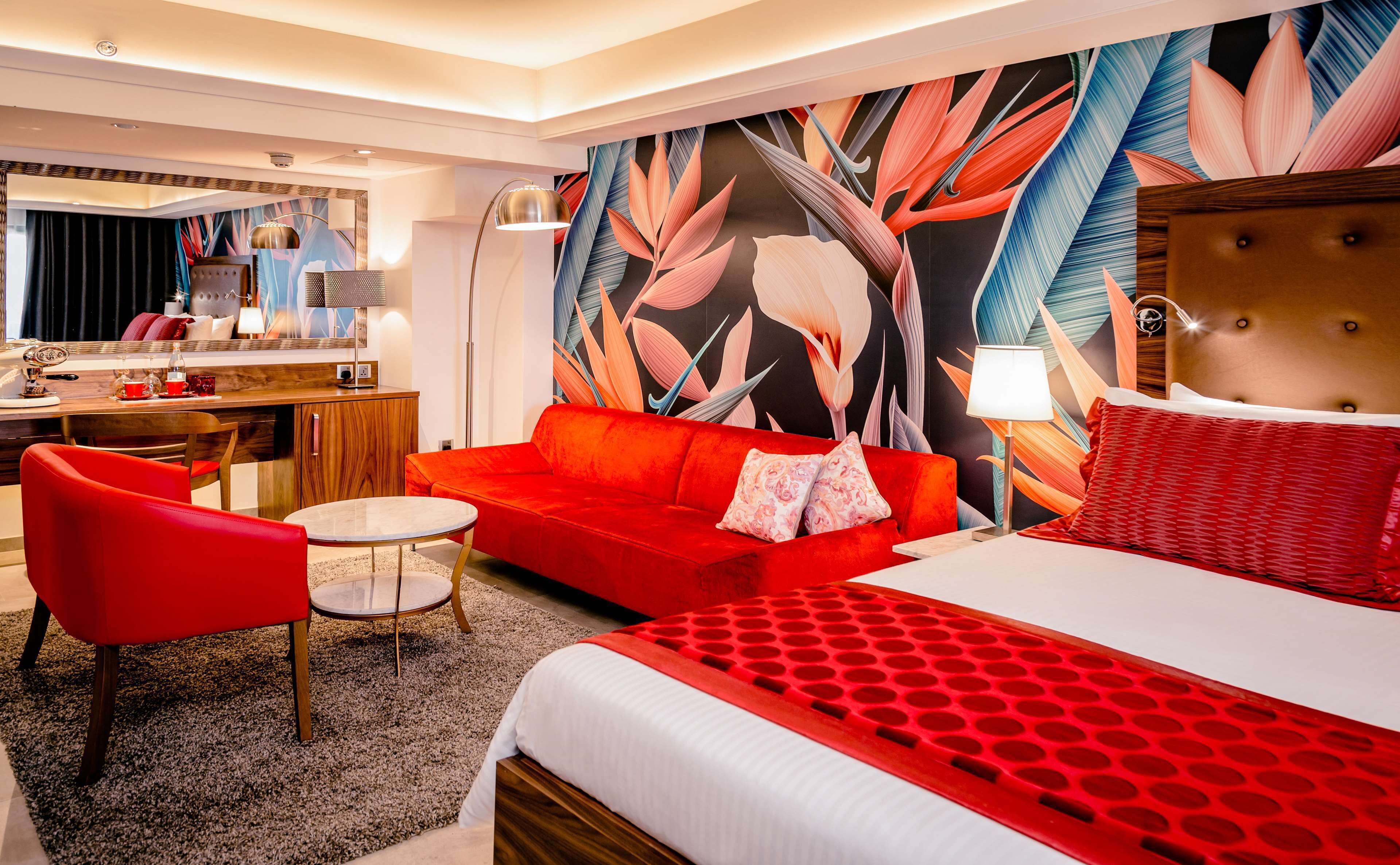 Habitación de hotel con sofá rojo, cama con colcha roja y blanca, una mesita redonda y una silla. Se observa un espejo grande, una lámpara y una pared con estampado de hojas tropicales.