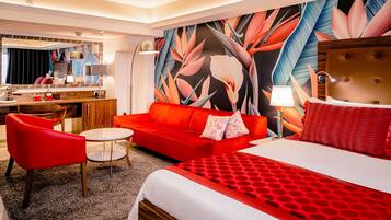 Habitación de hotel con sofá rojo, cama con colcha roja y blanca, una mesita redonda y una silla. Se observa un espejo grande, una lámpara y una pared con estampado de hojas tropicales.