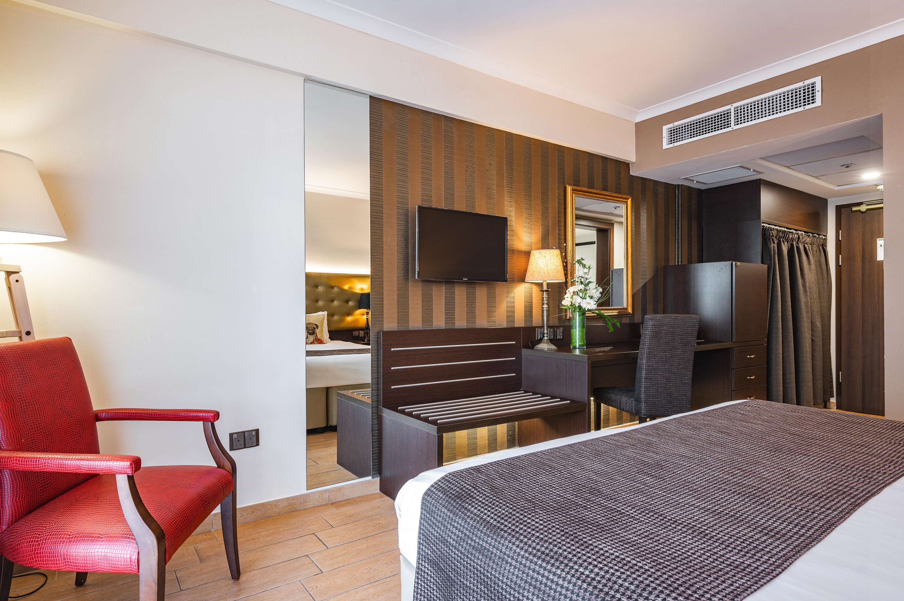 A hotel room with a bed, a red armchair, a desk with a chair, a TV, and a mirror.